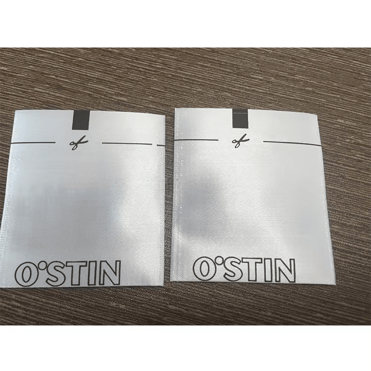 Două etichete albe pliate pentru articole de îmbrăcăminte textile RFID cu marca O STIN sunt plasate pe o masă pentru a arăta producția de etichete cusute personalizate.